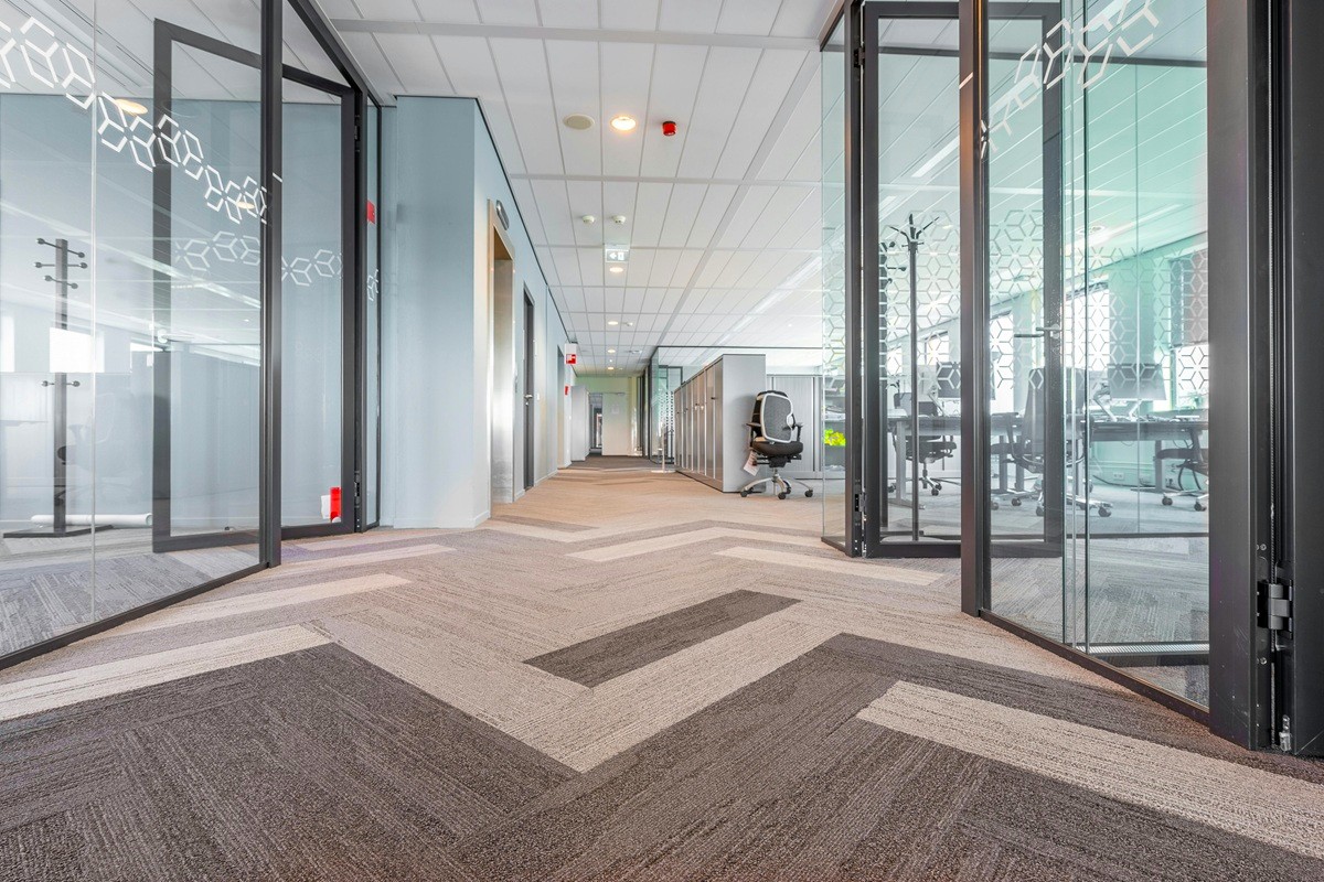 A modern office hallway with glass-walled rooms, chevron-patterned carpeting, and bright overhead lighting leading to workstations.