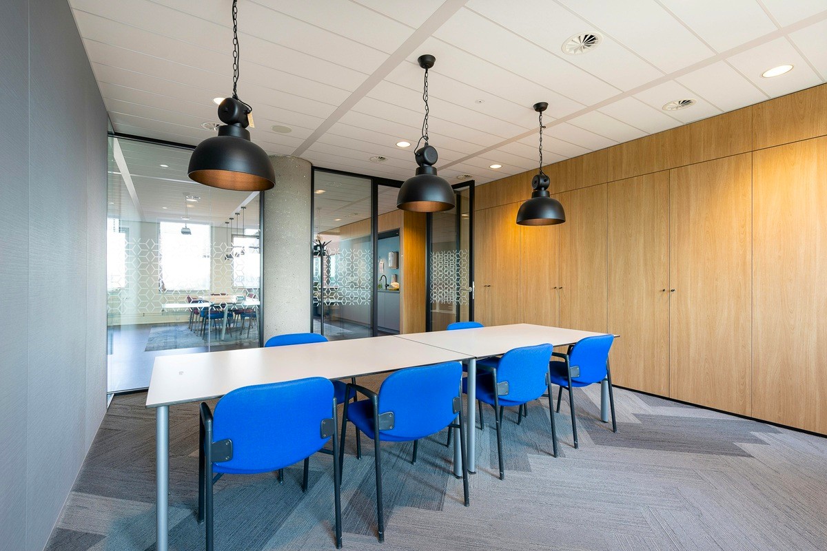 A small conference room with a long white table, blue chairs, and black pendant lighting, surrounded by wood-paneled walls and glass partitions.