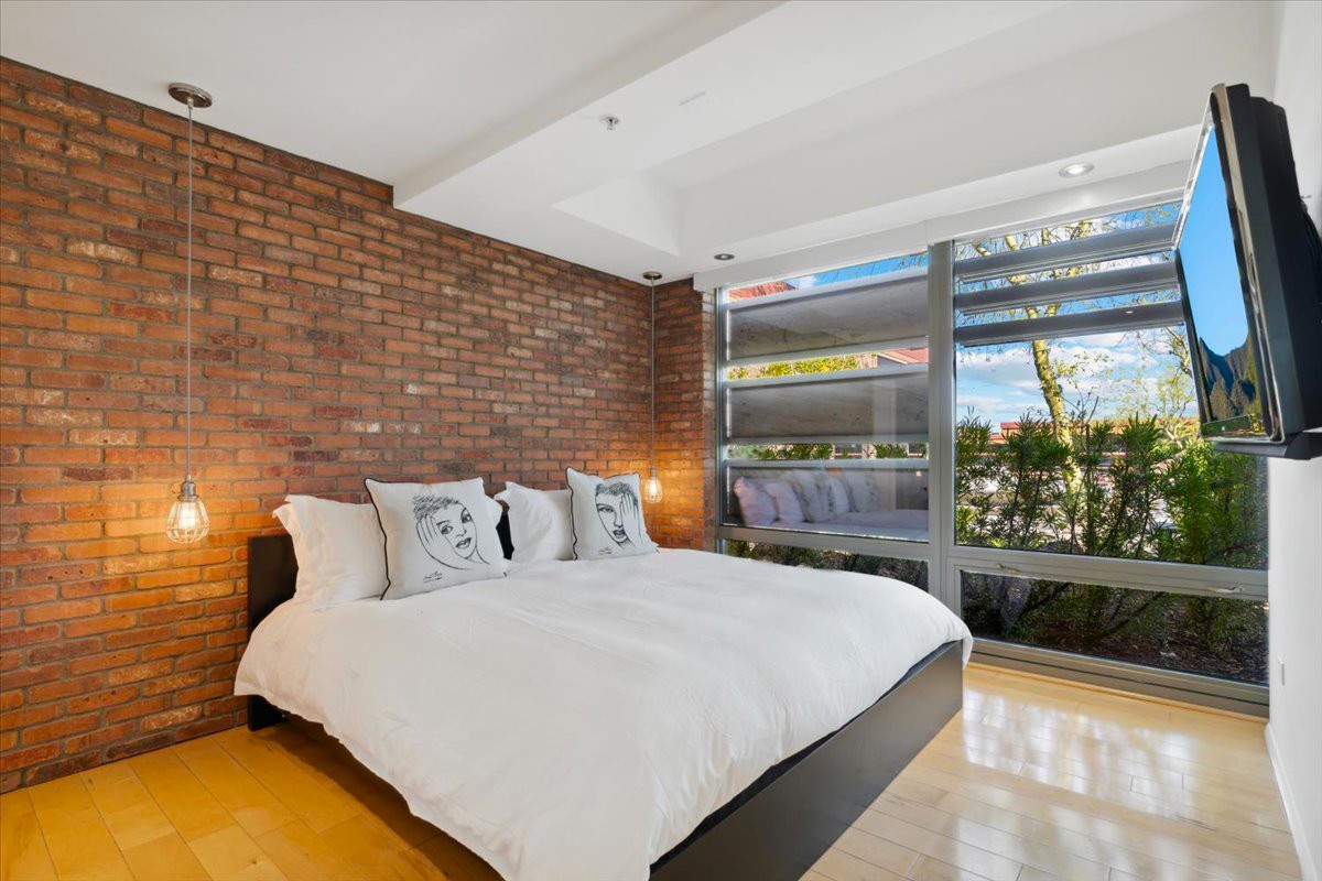 Optima Camelview condo bedroom with exposed brick wall, modern lighting, and large windows offering natural light and outdoor views of the development in Old Town.