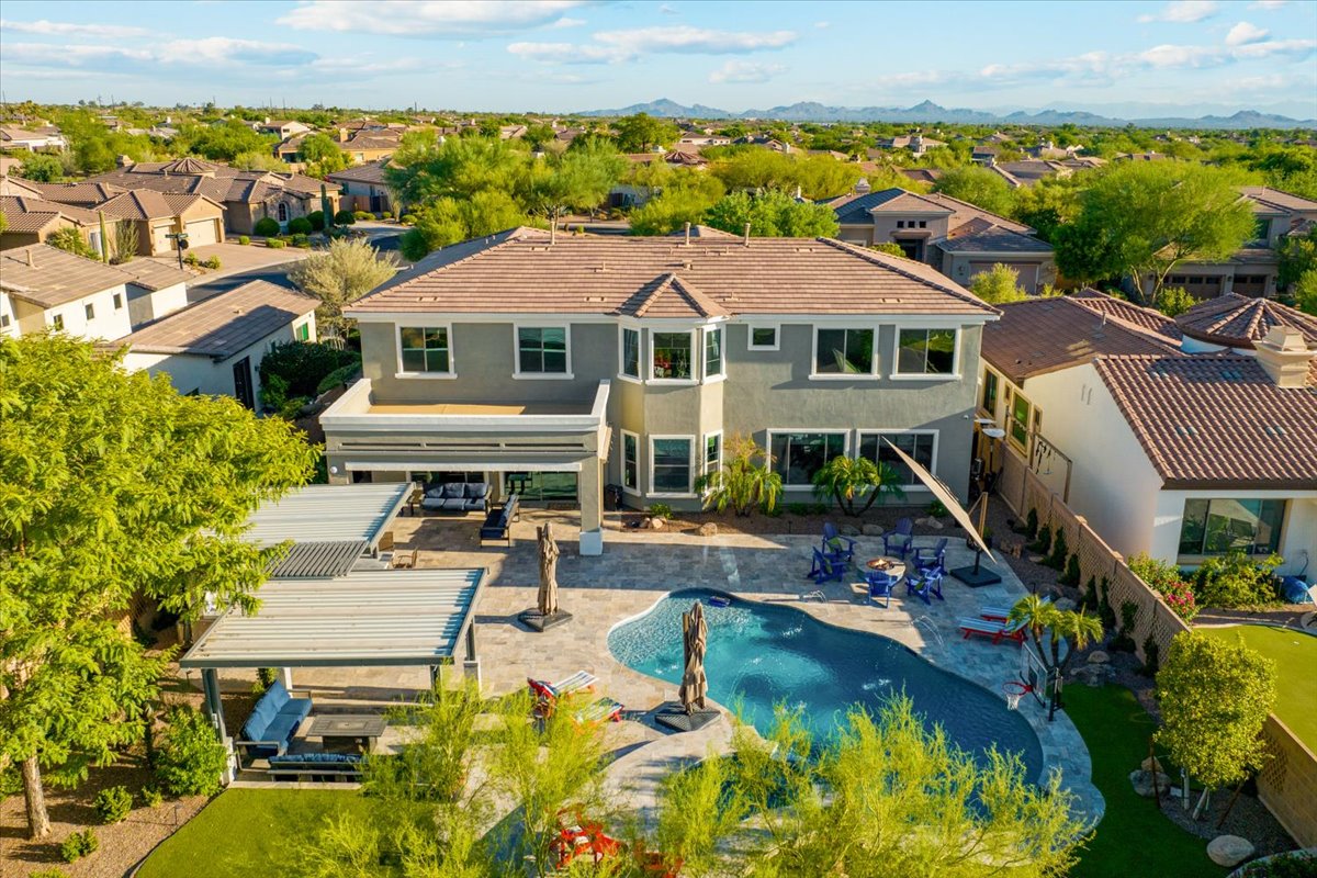 An aerial view of a spacious suburban home featuring a large backyard with a swimming pool, shaded patio areas, and landscaped surroundings in a quiet neighborhood.