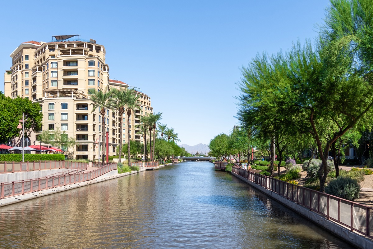 A scenic view of luxury condos along a peaceful canal, surrounded by lush greenery and palm trees under clear blue skies in Scottsdale.
