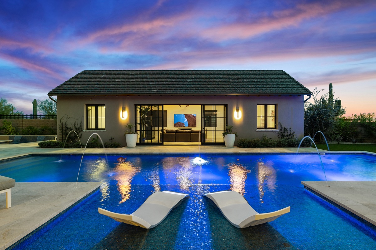 Backyard pool with water features, lounge chairs in the water, and a guest house illuminated at twilight.
