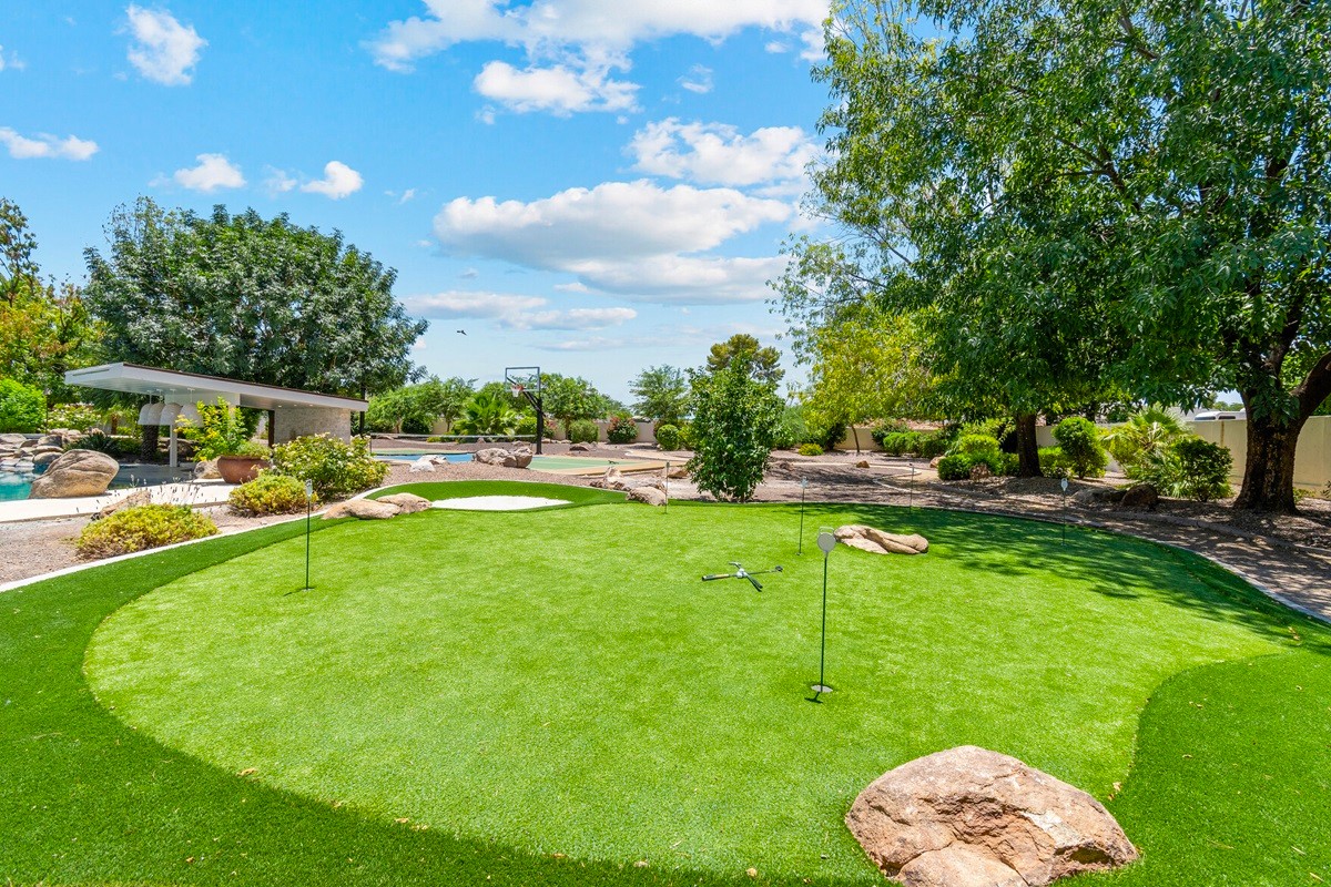 Outdoor putting green surrounded by desert landscaping, rocks, and trees, with a basketball hoop and pool visible in the background.