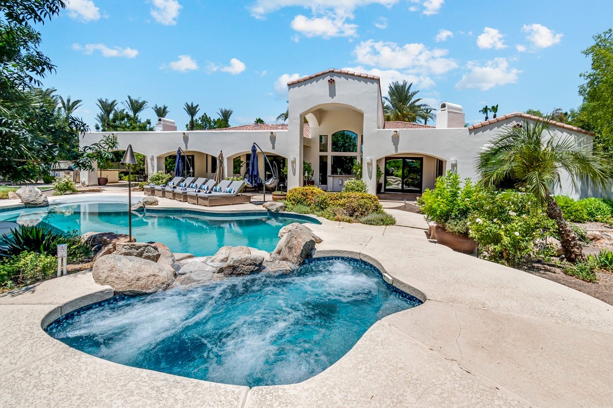 Resort-style pool and spa with lounge chairs, surrounded by lush landscaping and a beautiful white stucco home.