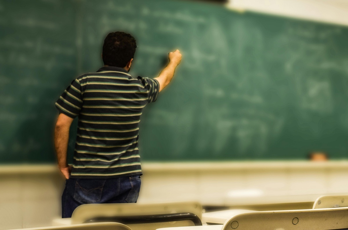 A teacher writing on a chalkboard in an empty classroom, viewed from behind.
