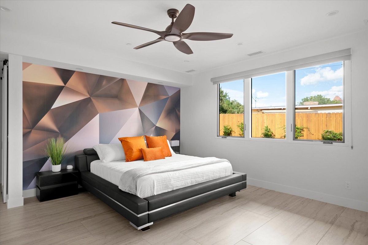 Modern bedroom with a geometric wall, white bedding, orange pillows, black platform bed, and large windows.