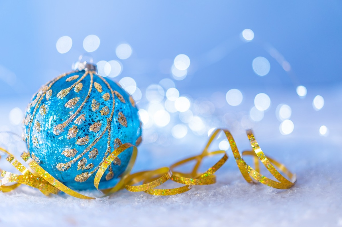 Blue ornament with gold details resting on snow, surrounded by golden ribbon and sparkling lights in the background. 