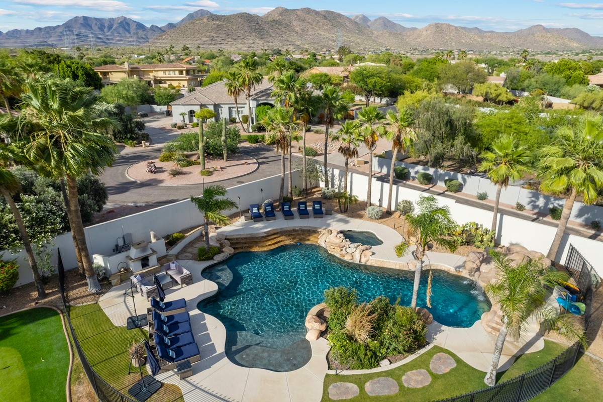 Aerial view of a backyard in a suburban neighborhood featuring a resort-style pool with a waterfall, lush landscaping, palm trees, and a mountain backdrop.