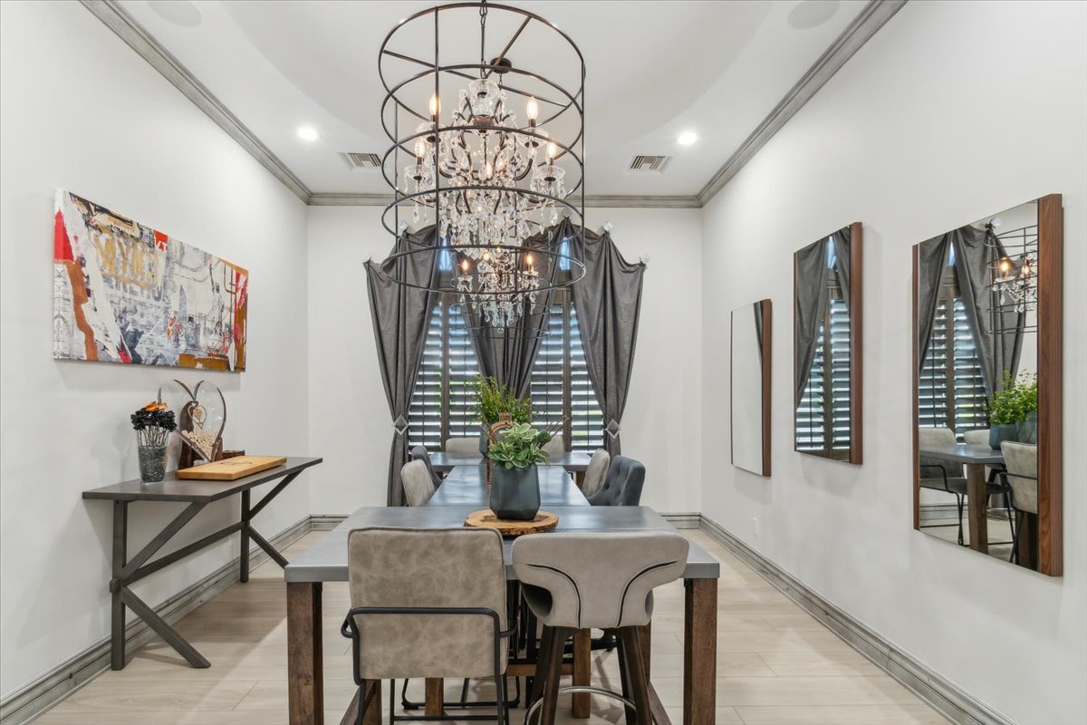 A modern dining space with a long table, upholstered chairs, and a striking chandelier inside a metal frame. Dark drapes, wall mirrors, and abstract art enhance the elegant decor.