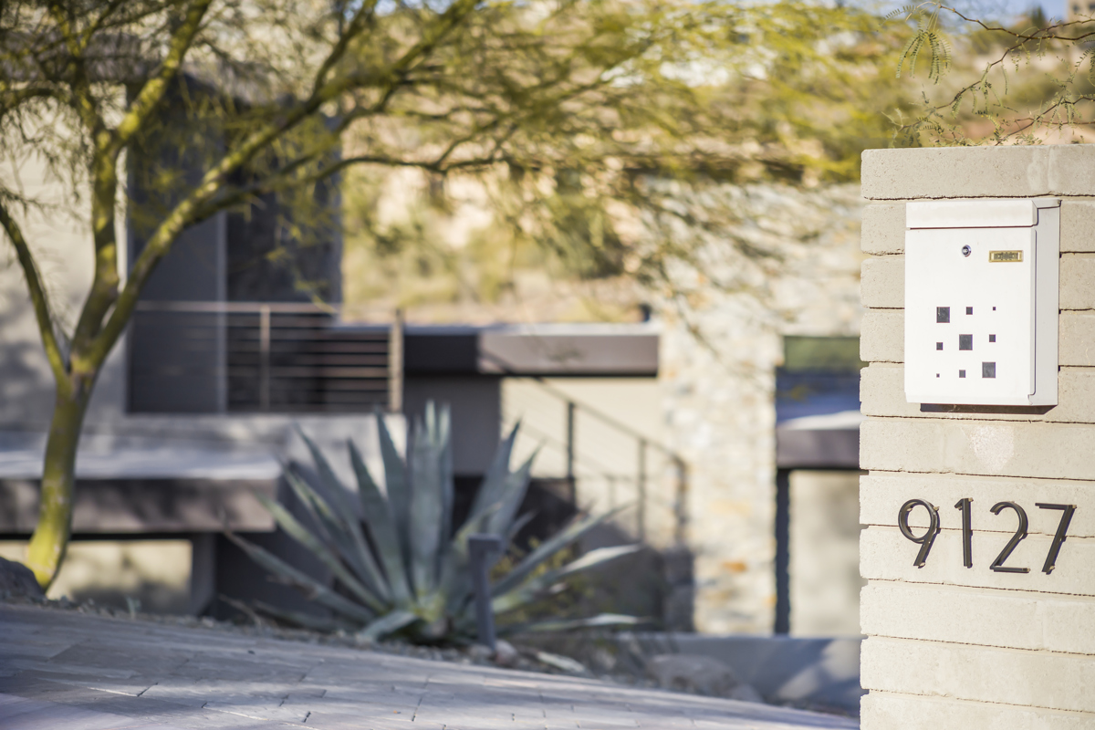 A close-up of a modern home’s entrance, featuring a stone wall with house numbers "9127" and a sleek mailbox. The background shows a desert-style landscape with agave plants.