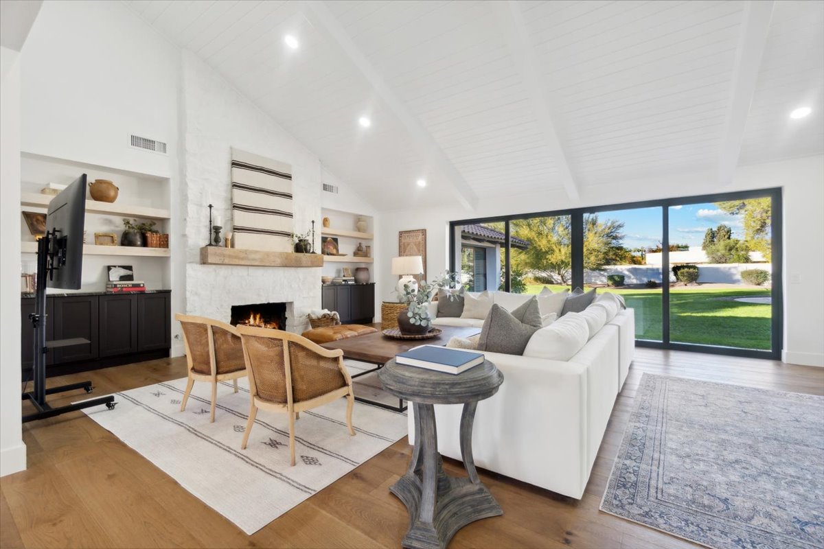 A bright and spacious living room with a vaulted white ceiling, a cozy fireplace, built-in shelves, and large sliding glass doors that open to a lush backyard.