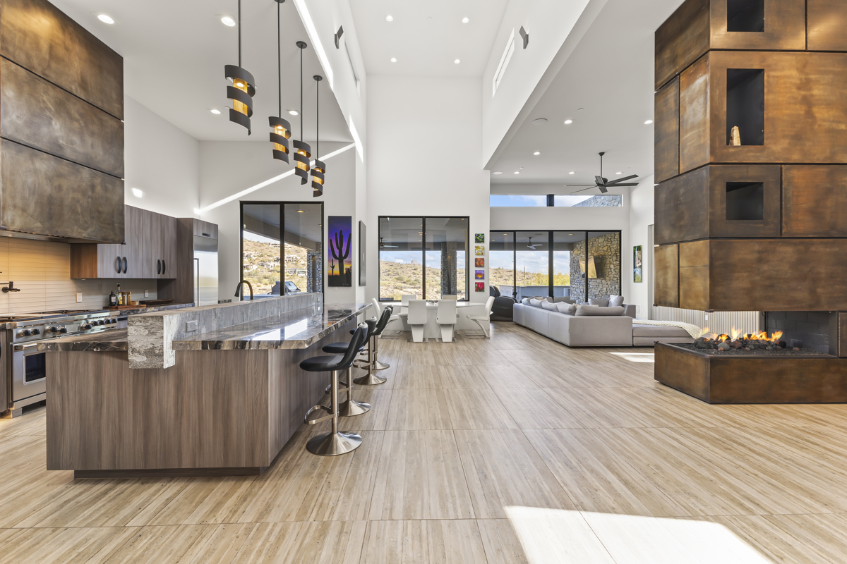A spacious modern living area with a sleek kitchen, bar seating, pendant lights, a statement fireplace, and floor-to-ceiling windows showcasing a desert view.