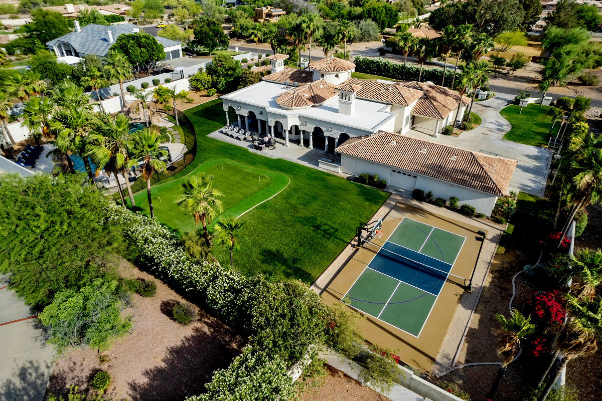 An aerial view of a Mediterranean-style estate with a lush lawn, a pool, a pickleball court, and palm trees surrounding the property.