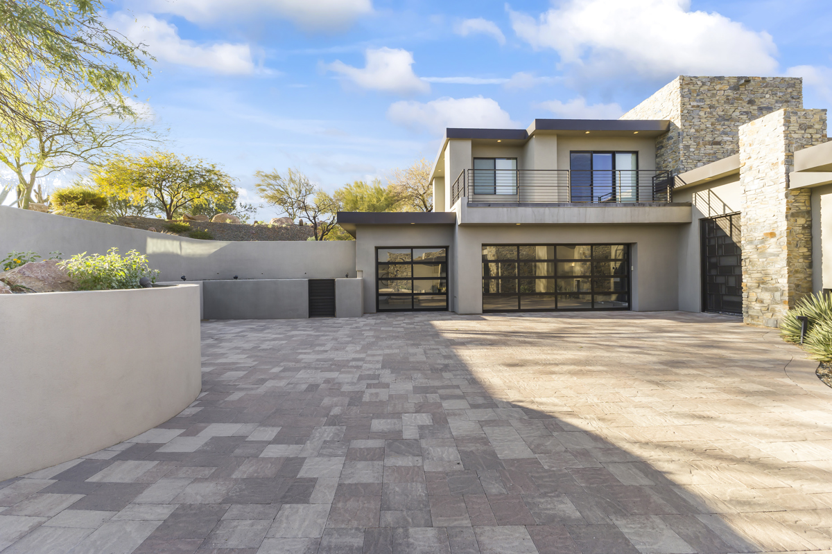 A modern luxury home with stone accents, large glass doors, and a spacious paved driveway under a bright blue sky.
