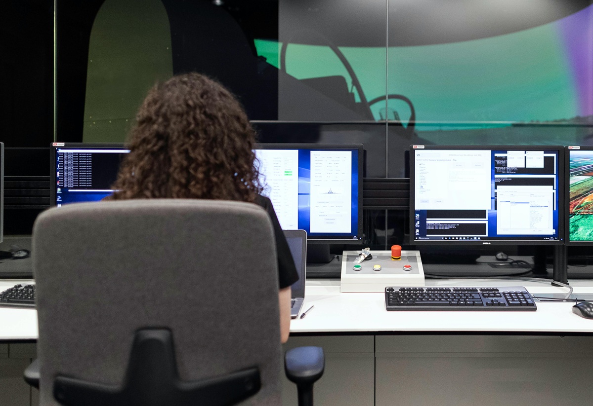 A woman with curly hair sits at a workstation with multiple monitors displaying data and controls.