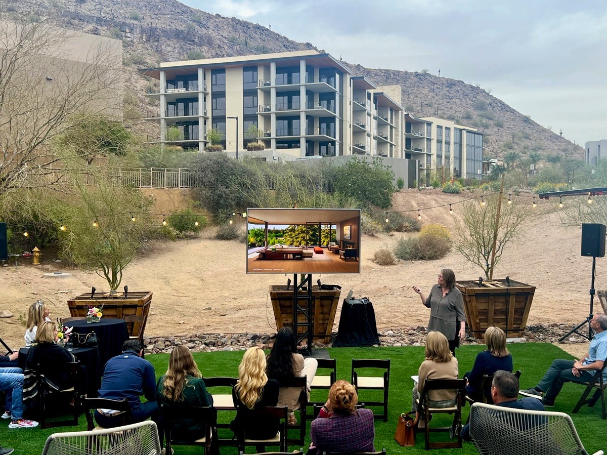 Woman giving a presentation for Summit by Olsen Kundig to attendees seated outdoors, framed by desert scenery and contemporary buildings.