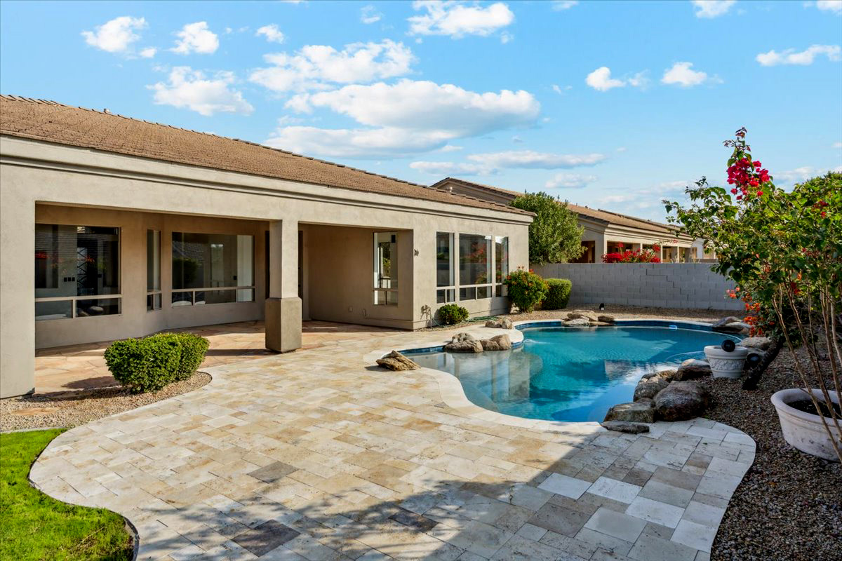 Private backyard featuring a curved swimming pool, stone paver patio, mature landscaping, and a covered patio area along the home.