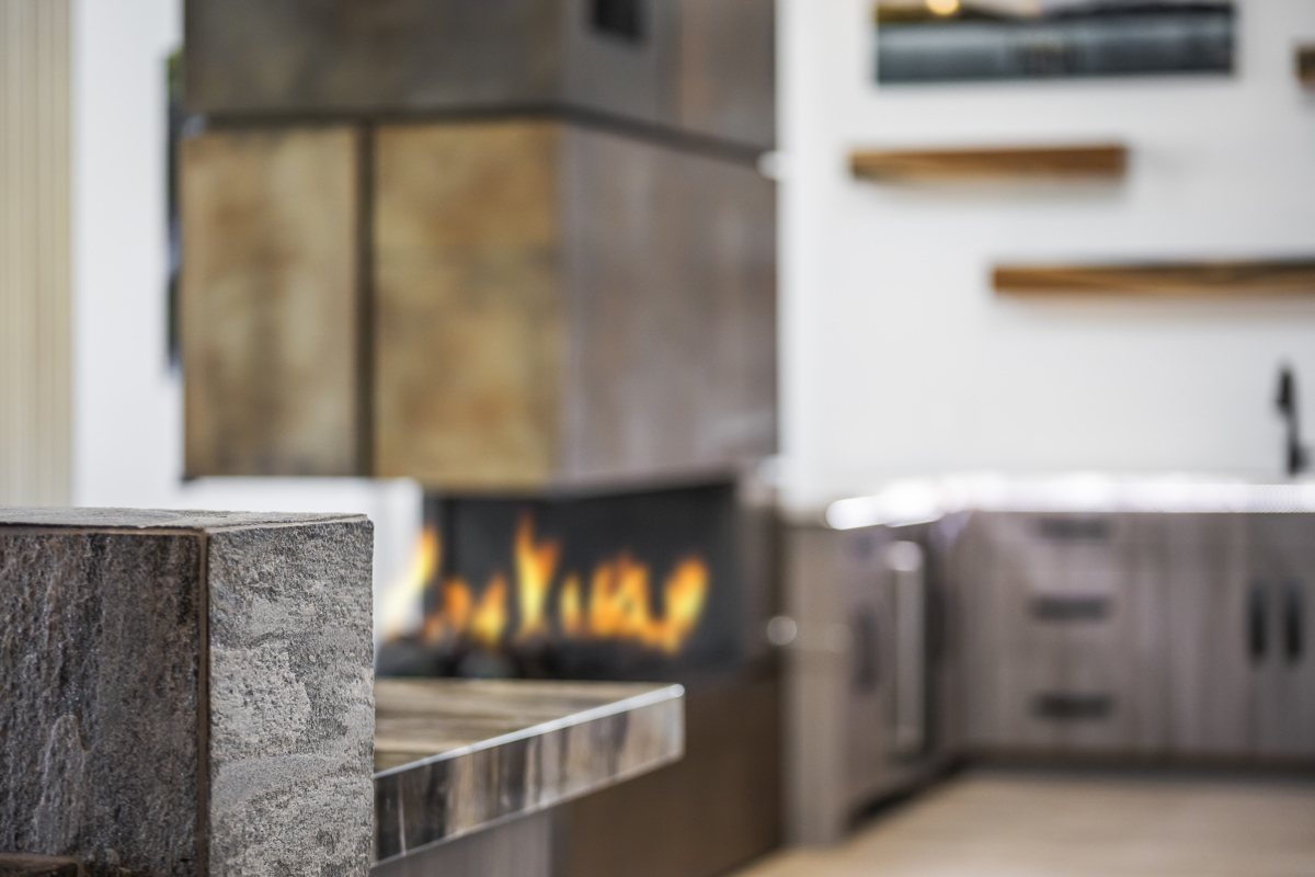 A modern kitchen with a stone countertop in the foreground and a sleek, double-sided fireplace with flames visible in the middle. The background features contemporary cabinetry and shelving.