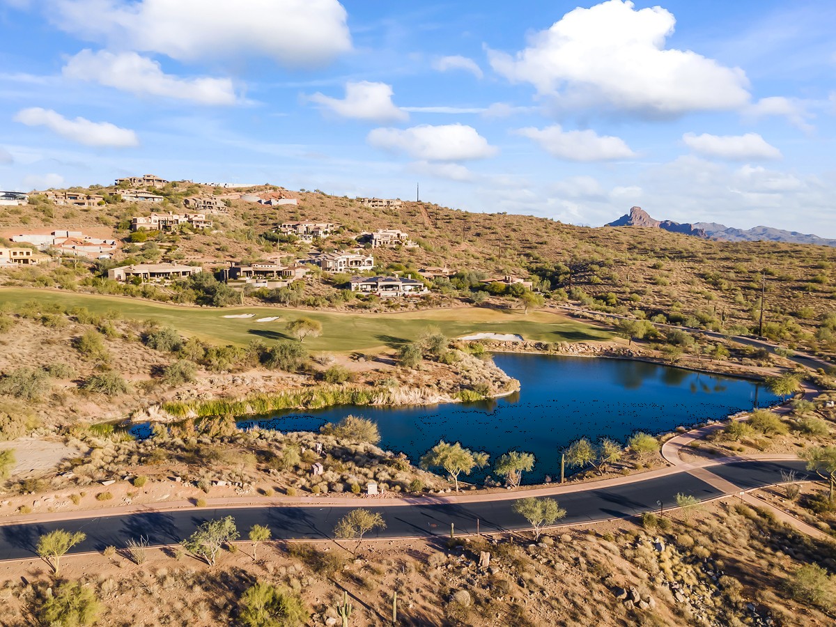 A scenic desert golf course community with luxury homes, a lake, and mountain views under a bright blue sky.