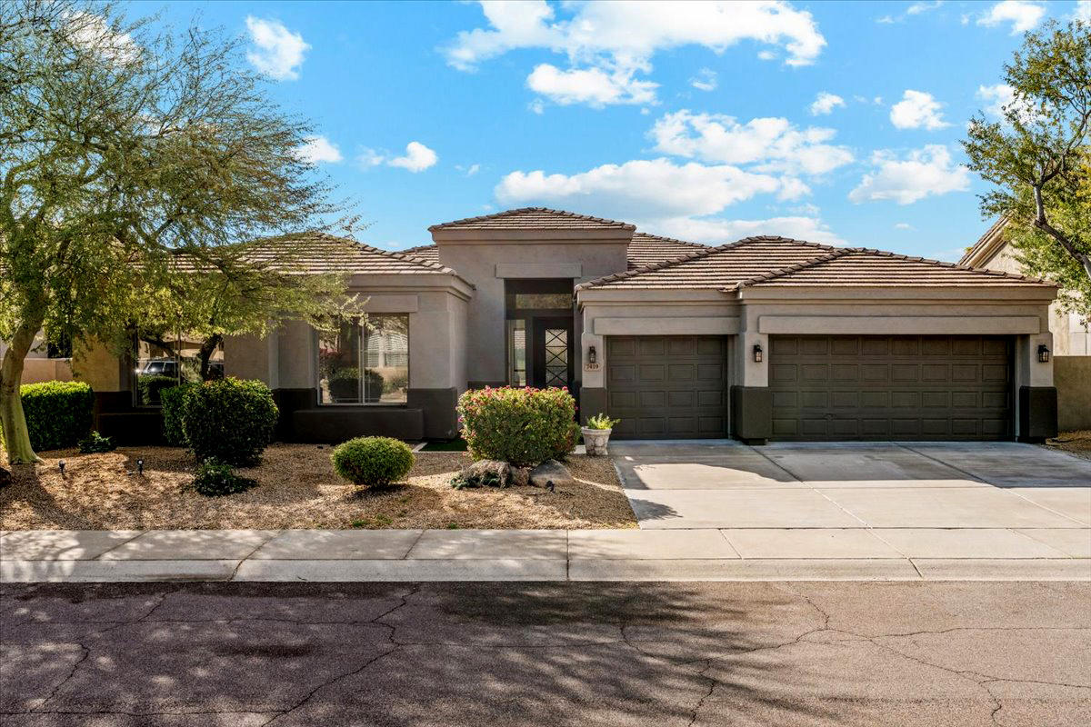 Single-story home with a three-car garage, flat rooflines, desert landscaping, and a stucco exterior in neutral tones under a blue sky.