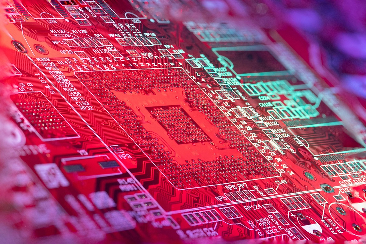 A colorful red circuit board illuminated by pink and blue lighting, showcasing fine electronic traces and component slots.