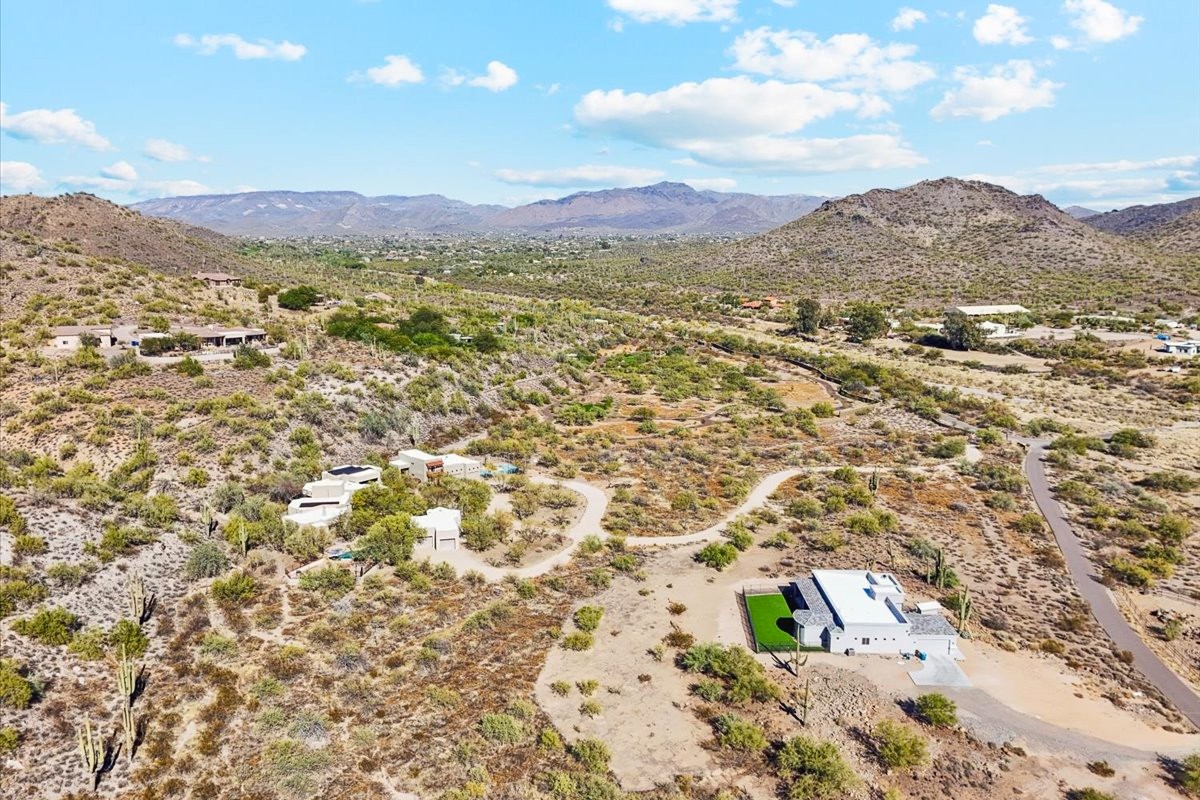 Aerial view of a desert neighborhood in Scottsdale, Arizona, with scattered homes, winding roads, and rugged mountains in the background under a bright blue sky.