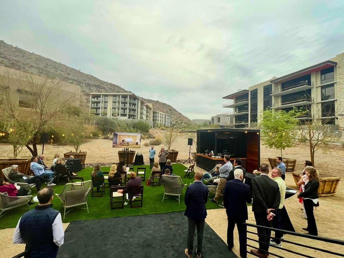 Outdoor presentation event with audience seated on grass, surrounded by desert landscape and modern buildings.