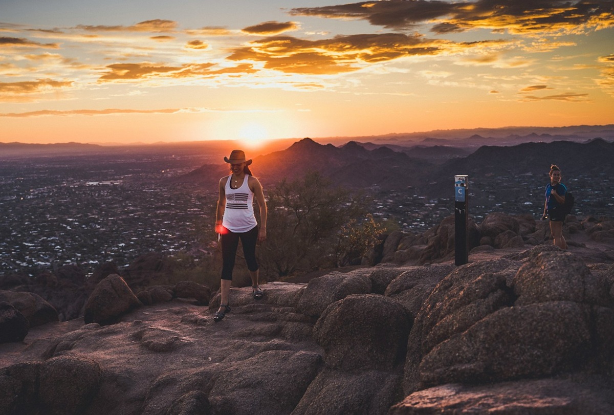 A hiker walks near the summit of Camelback Mountain in Phoenix, Arizona, with a warm desert sunset glowing behind the ridgeline.