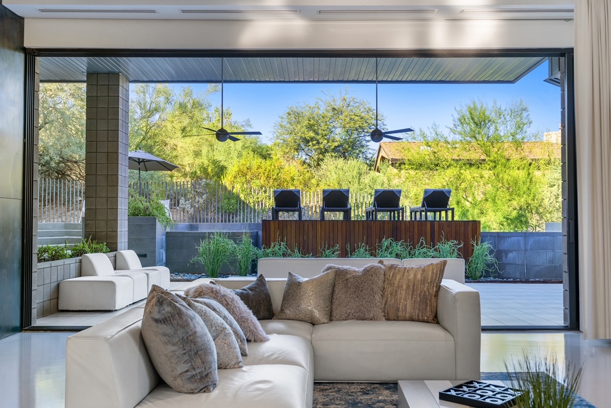 View from indoor living space looking out to a shaded patio with ceiling fans, modern outdoor furniture, and lush greenery behind a wooden privacy wall.