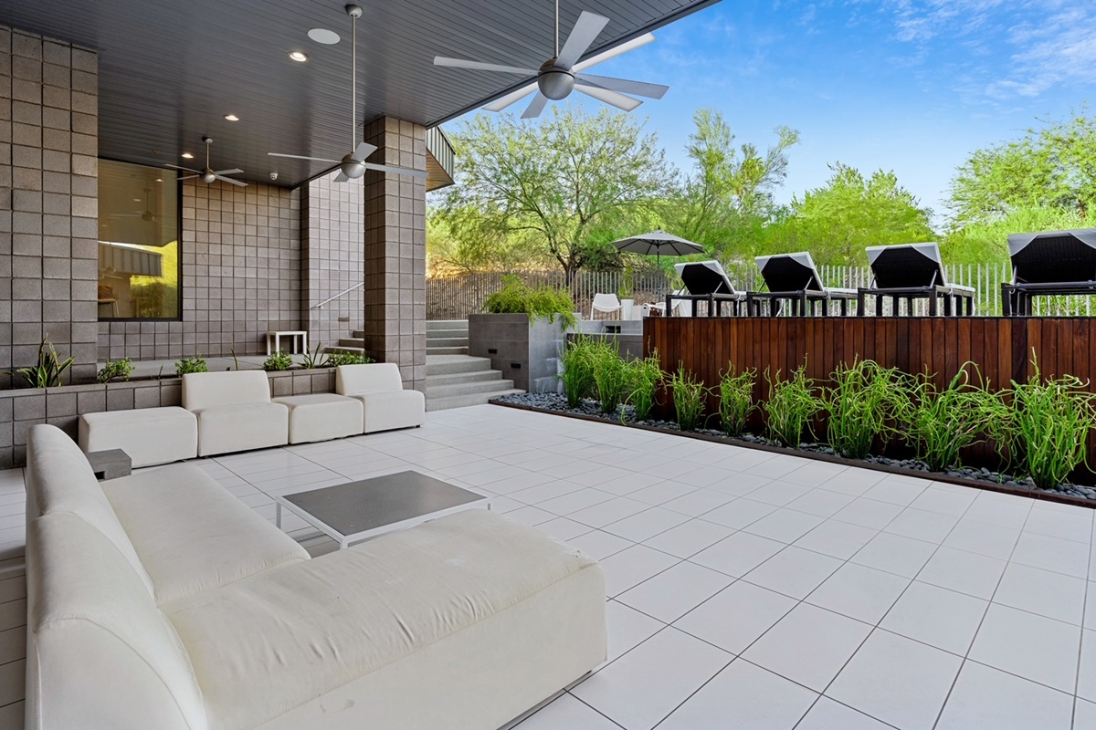 Covered outdoor patio with white modern seating, tile flooring, ceiling fans, and a raised wood-deck bar area lined with black lounge chairs and green landscaping.