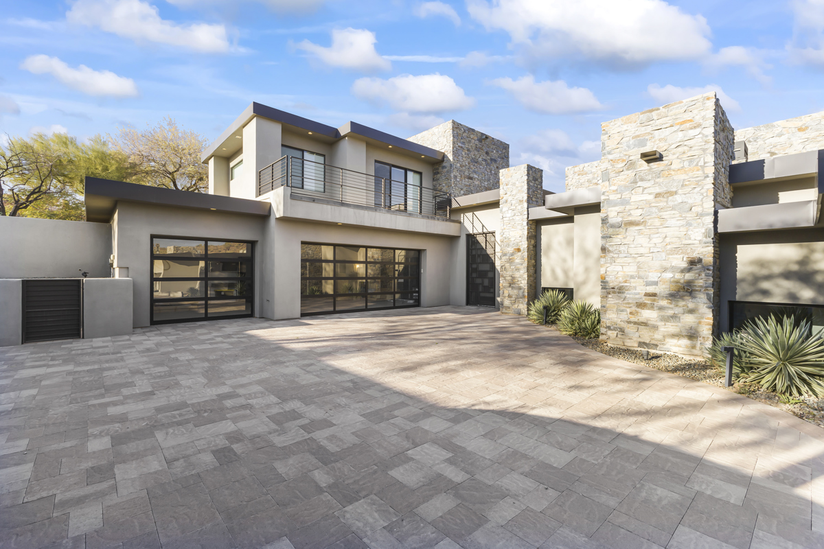 Modern desert-style home in Greater Phoenix with stone accents, large glass garage doors, and a spacious paver driveway under a bright blue sky.