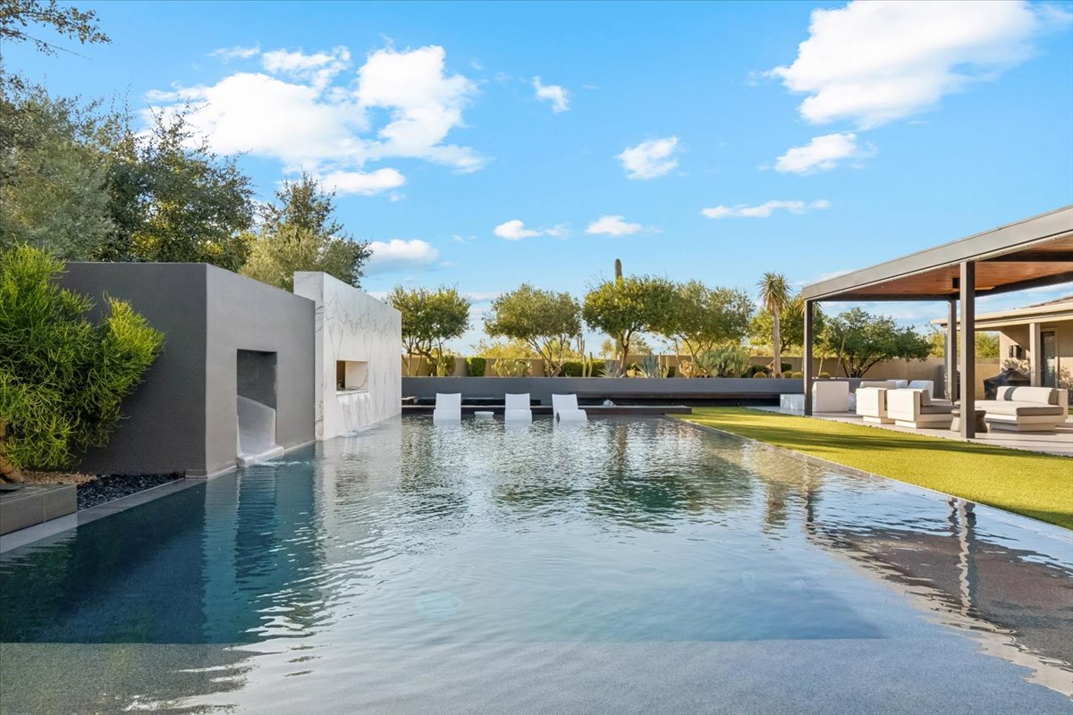 Modern backyard with infinity pool, water feature, in-pool lounge chairs, and surrounding greenery under a clear blue sky.