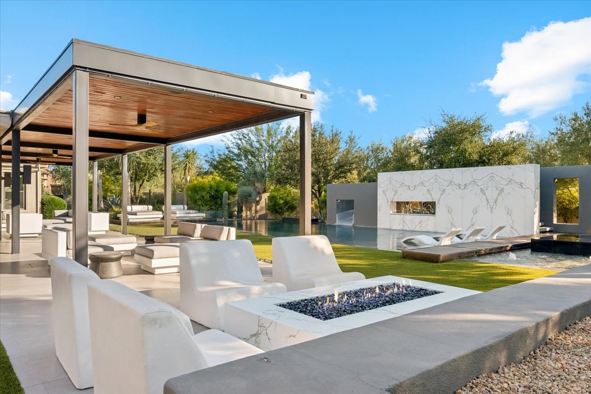 Outdoor lounge with white seating, marble fire pit, pergola, and pool area with sun loungers and modern landscaping.