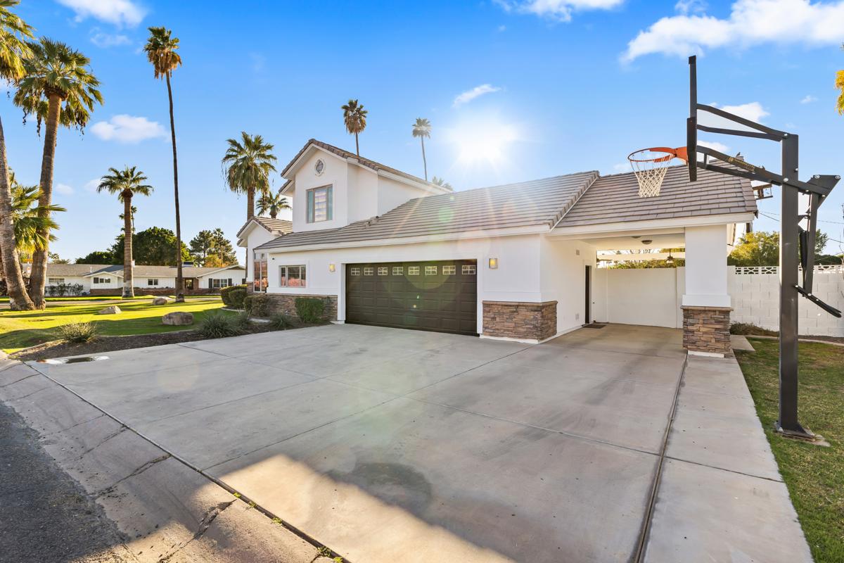 Two-story suburban house with white walls, brown garage doors, and stone trim, surrounded by palm trees and a spacious driveway under a sunny sky.
