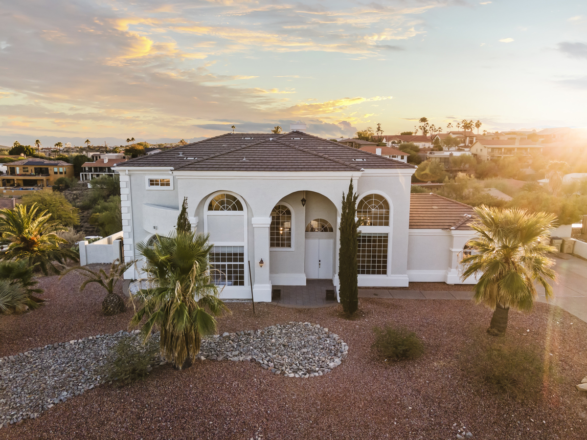 A large two-story desert home with arched windows, palm trees, and a rocky front yard at sunset.