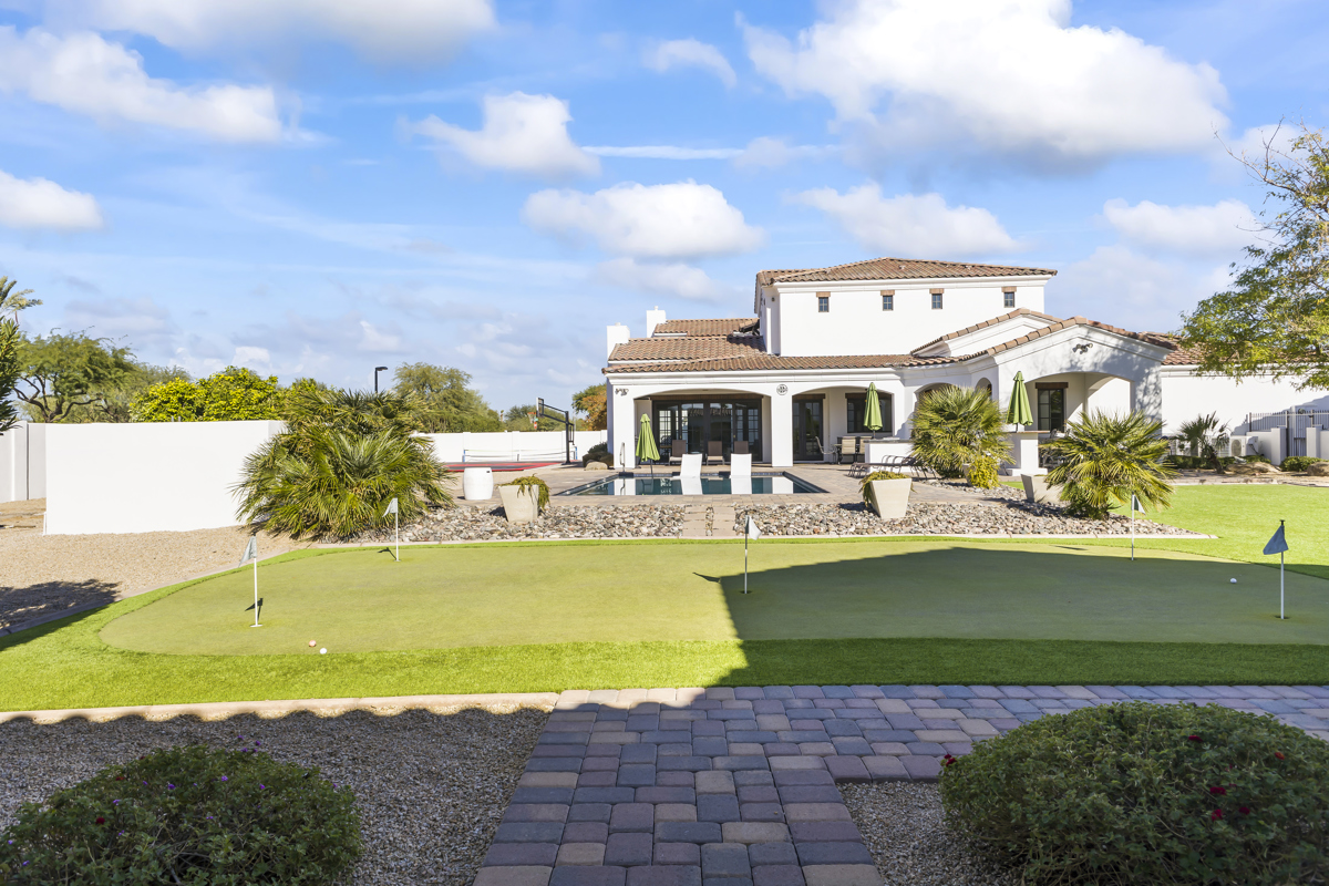 A backyard view of a white villa with a pool, patio umbrellas, and a putting green in the foreground.