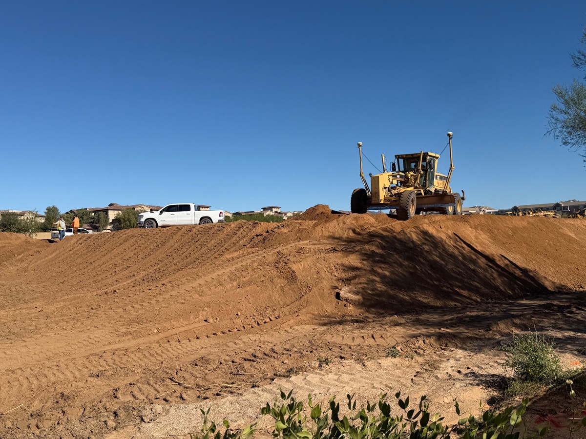 Heavy grader leveling soil on an active construction site with trucks parked nearby.