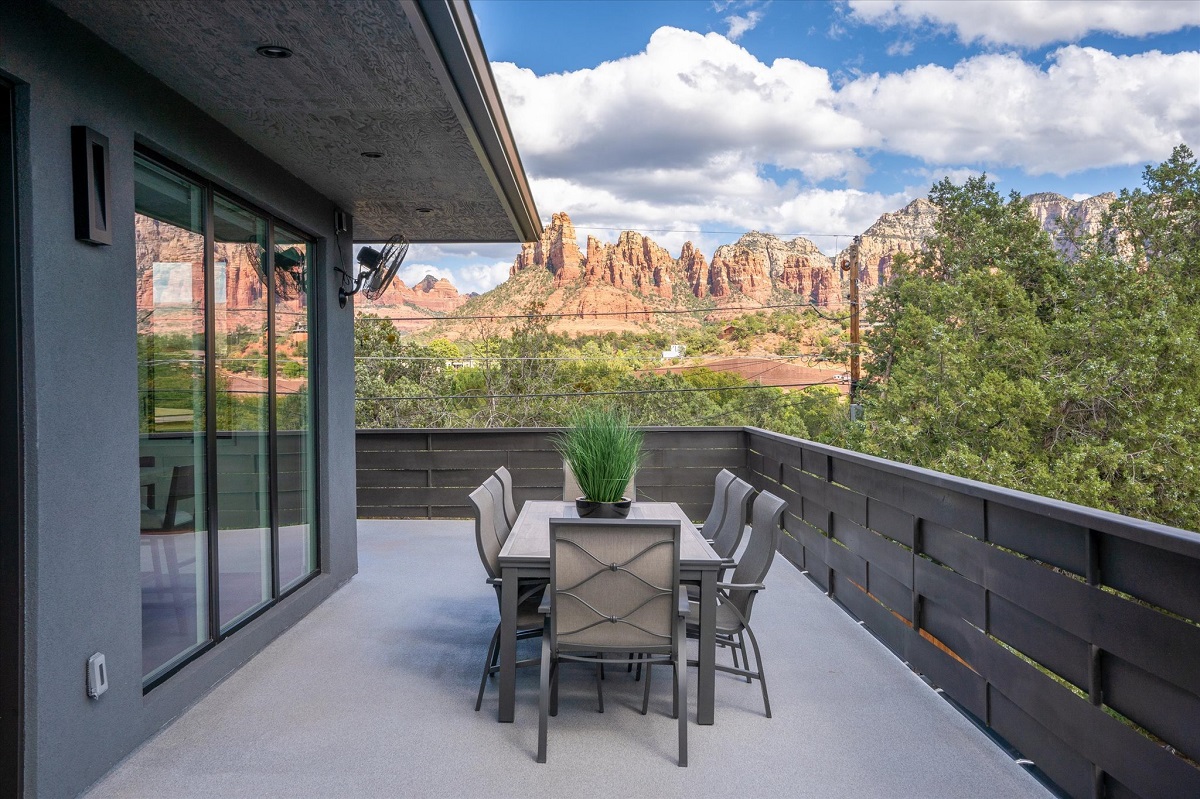 Modern balcony with outdoor dining set overlooking red rock cliffs and green trees under a blue sky.