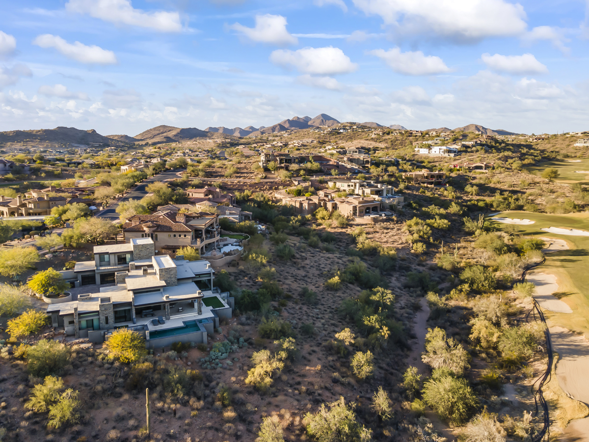 Aerial view of a desert golf course with green fairways, a small lake, and mountain homes nearby.