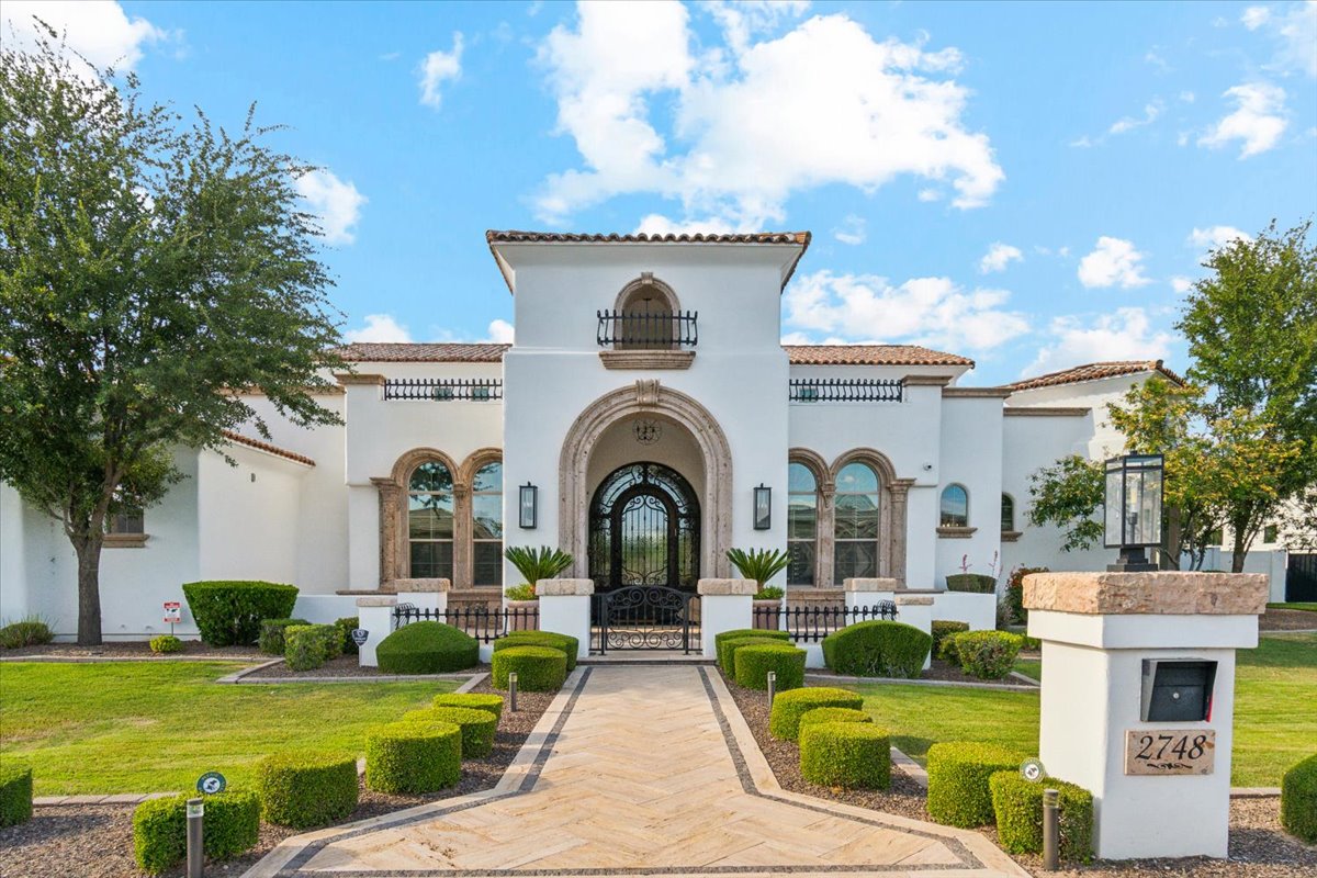 Large white stucco house with arched entryway, manicured landscaping, and a paved front walkway.