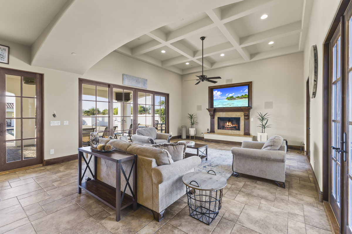 Bright, spacious living room with coffered ceiling, tile floors, fireplace with TV above, and large glass doors opening to a covered patio.