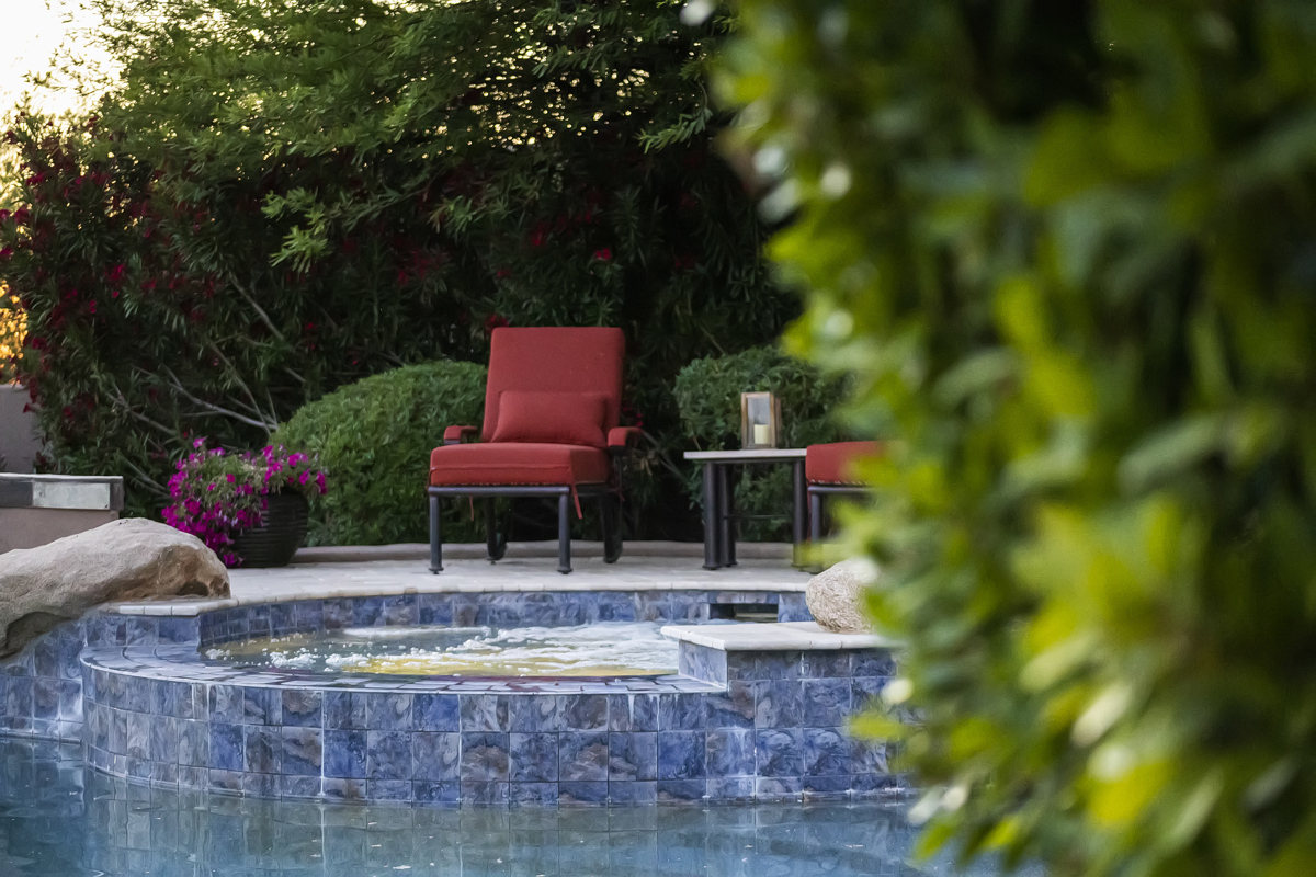 Backyard spa with blue tile, red patio chairs, greenery, & poolside seating.