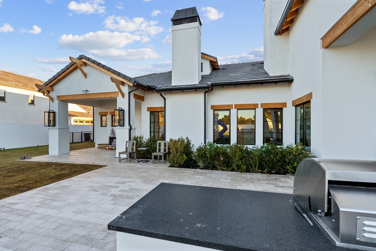 A photo of a luxury home patio with white stucco walls, wood trim, a covered seating area, and an outdoor grill.