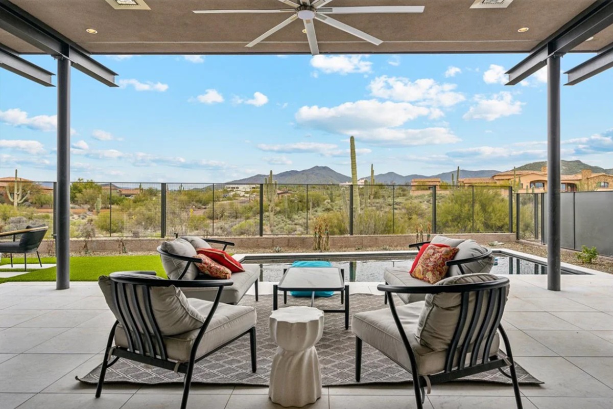 A photo of a covered patio with outdoor seating, a pool, desert landscaping, and mountain views.