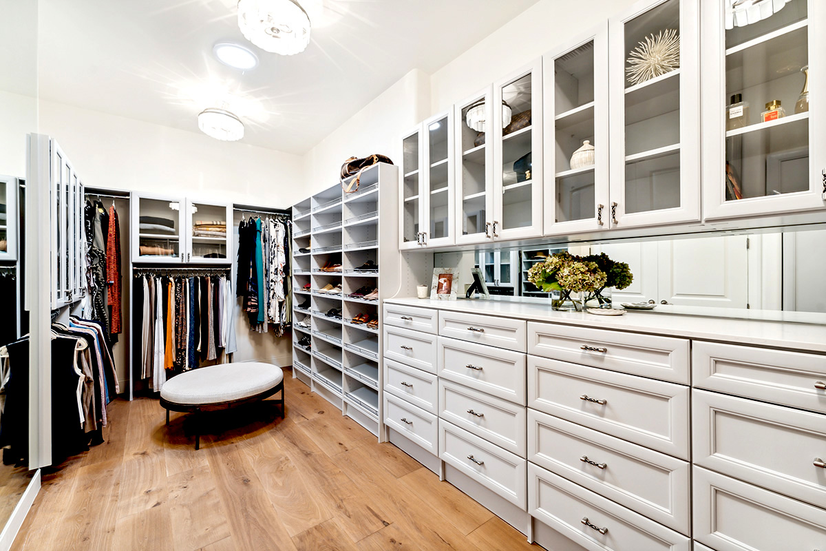 Luxury designer walk-in closet with custom white cabinetry, glass-front storage & hardwood floors.