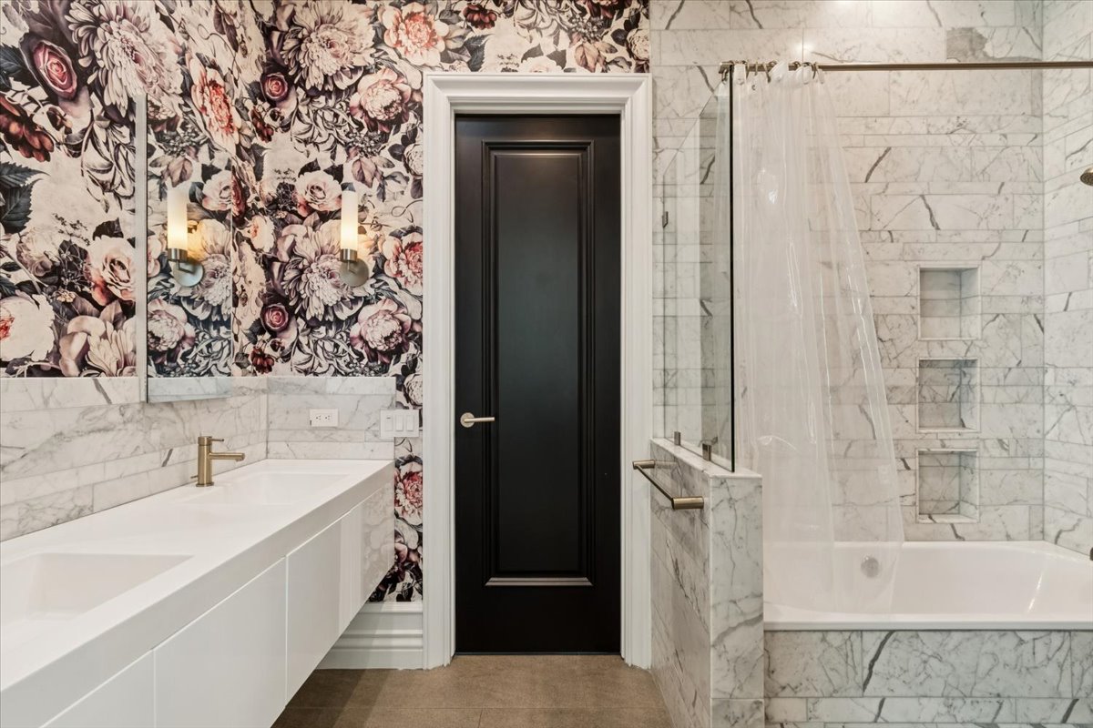 Photo of a designer bathroom with floral wallpaper, a white double vanity, brass fixtures, and a marble bathtub surround beside a black door.