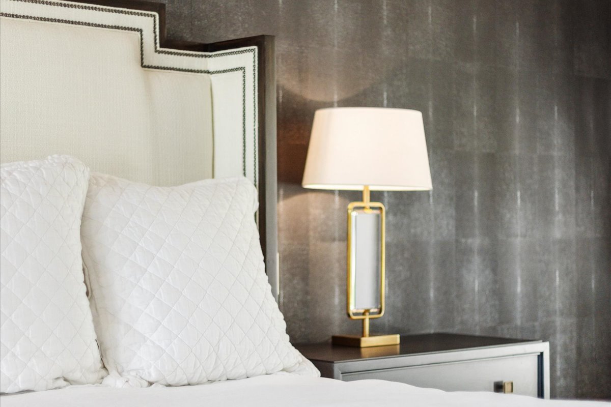 Photo of a luxury bedroom featuring a white upholstered headboard, quilted pillows, textured gray walls, and a gold-accented bedside lamp.