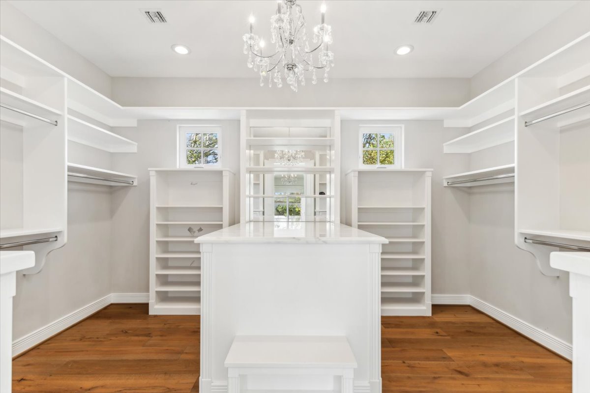 Spacious luxury walk-in closet with white built-in shelving, a central island, crystal chandelier, and wood flooring.