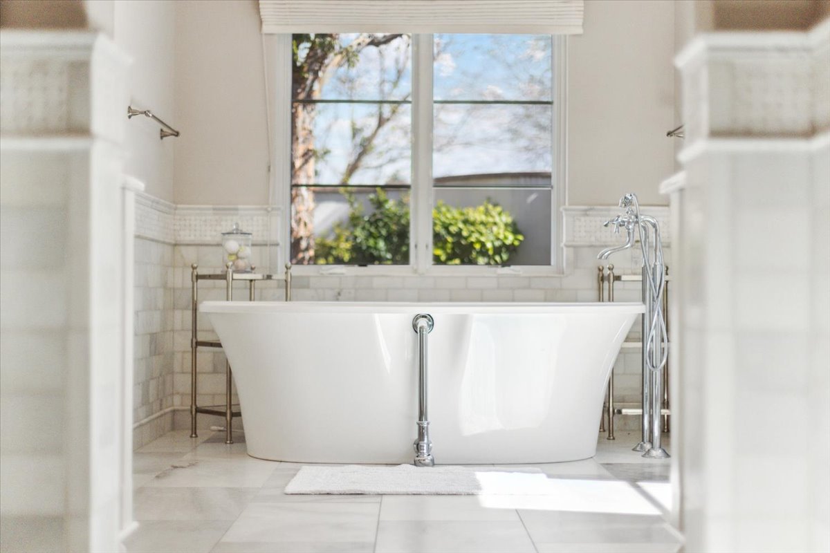 Elegant freestanding white bathtub with polished chrome fixtures, tile flooring, and a large window overlooking greenery.