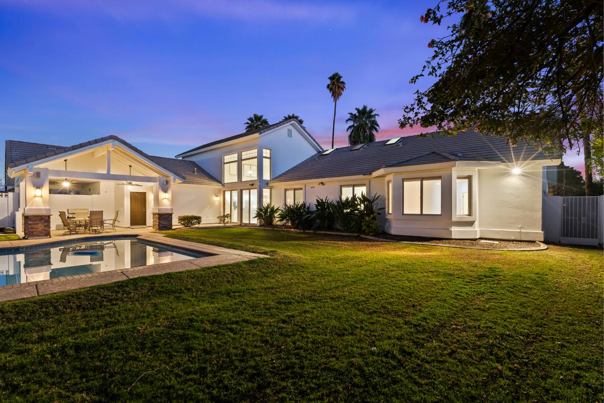 Arizona home featuring a spacious backyard, swimming pool, covered patio, and illuminated exterior at dusk.